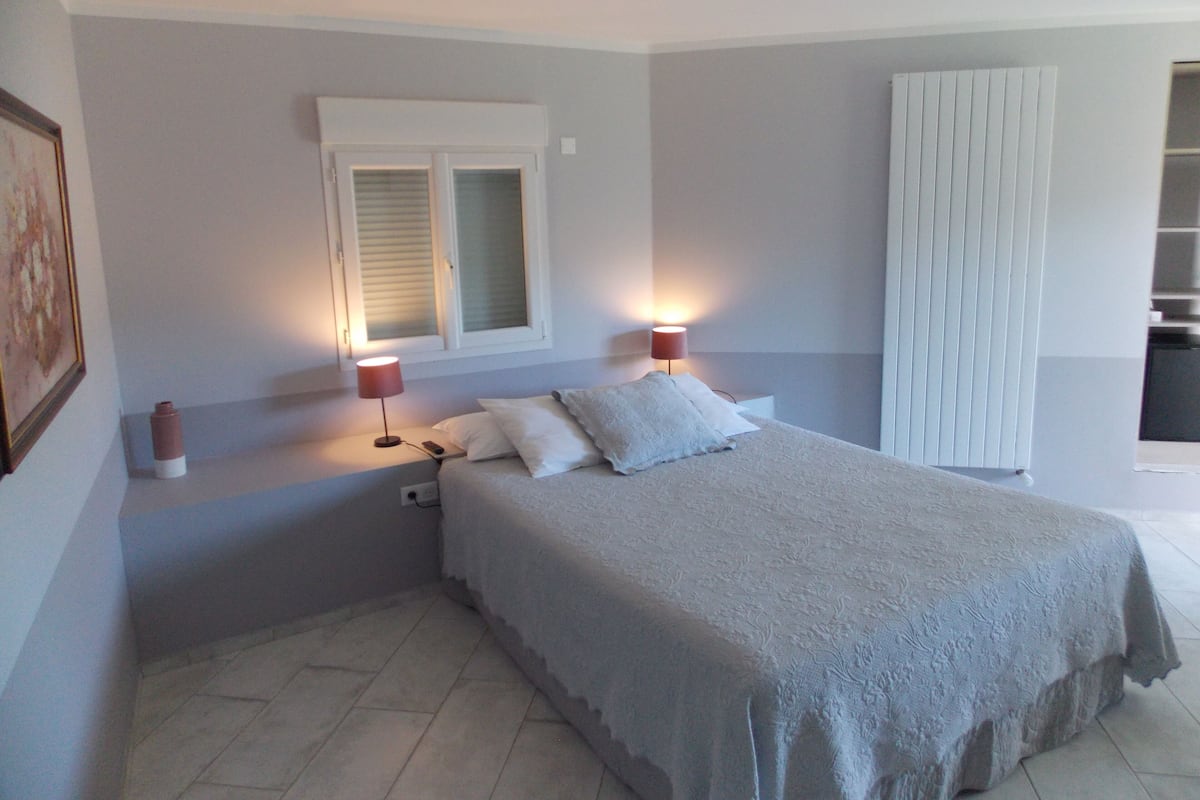 Quarto com uma cama, duas luminárias de cabeceira, uma janela com persianas e uma porta para o banheiro.