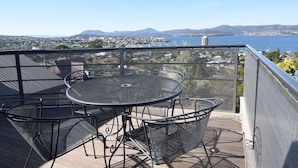A balcony with a round table and chairs, overlooking a coastal cityscape with mountains in the distance. - StudioAt10 (Sandy Bay)