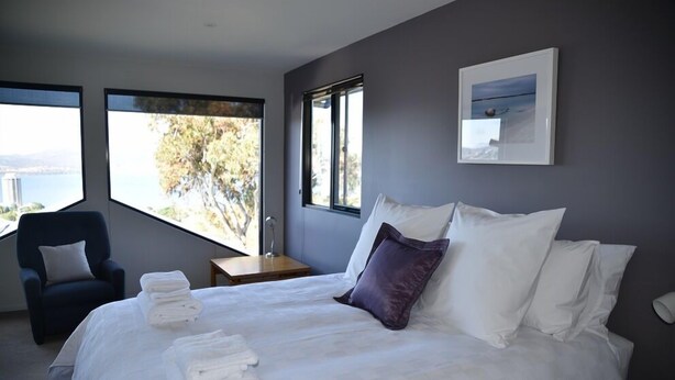 A modern hotel room with a large bed, a chair, and a view of a body of water through large windows. - StudioAt10 (Sandy Bay)