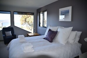 A modern hotel room with a large bed, a chair, and a view of a body of water through large windows. - StudioAt10 (Sandy Bay)