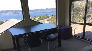A room with a large window offering a view of a body of water and distant hills. There is a black desk with two chairs and a purple ottoman. - StudioAt10 (Sandy Bay)