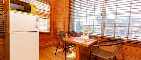 Interior de una cabaña de madera con un refrigerador blanco, una mesa de comedor de madera, dos sillas de mimbre y una ventana con persianas.