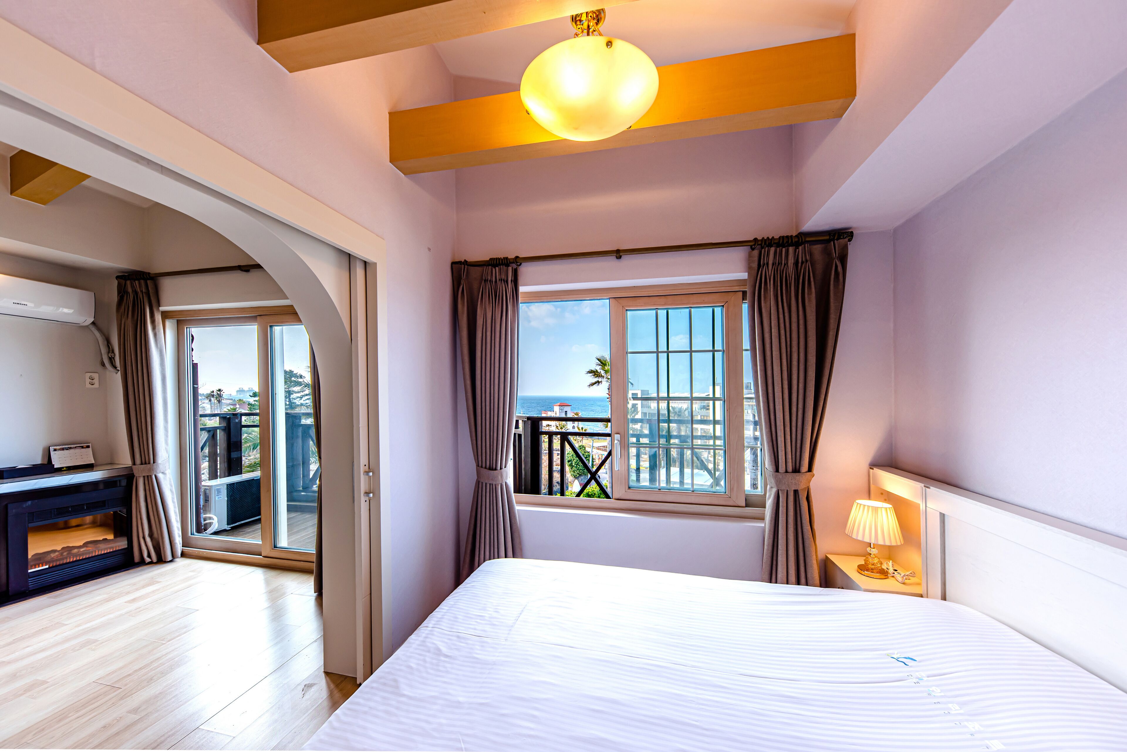 A bedroom with a large bed, a nightstand with a lamp, a window with curtains, and a view of the outdoors.