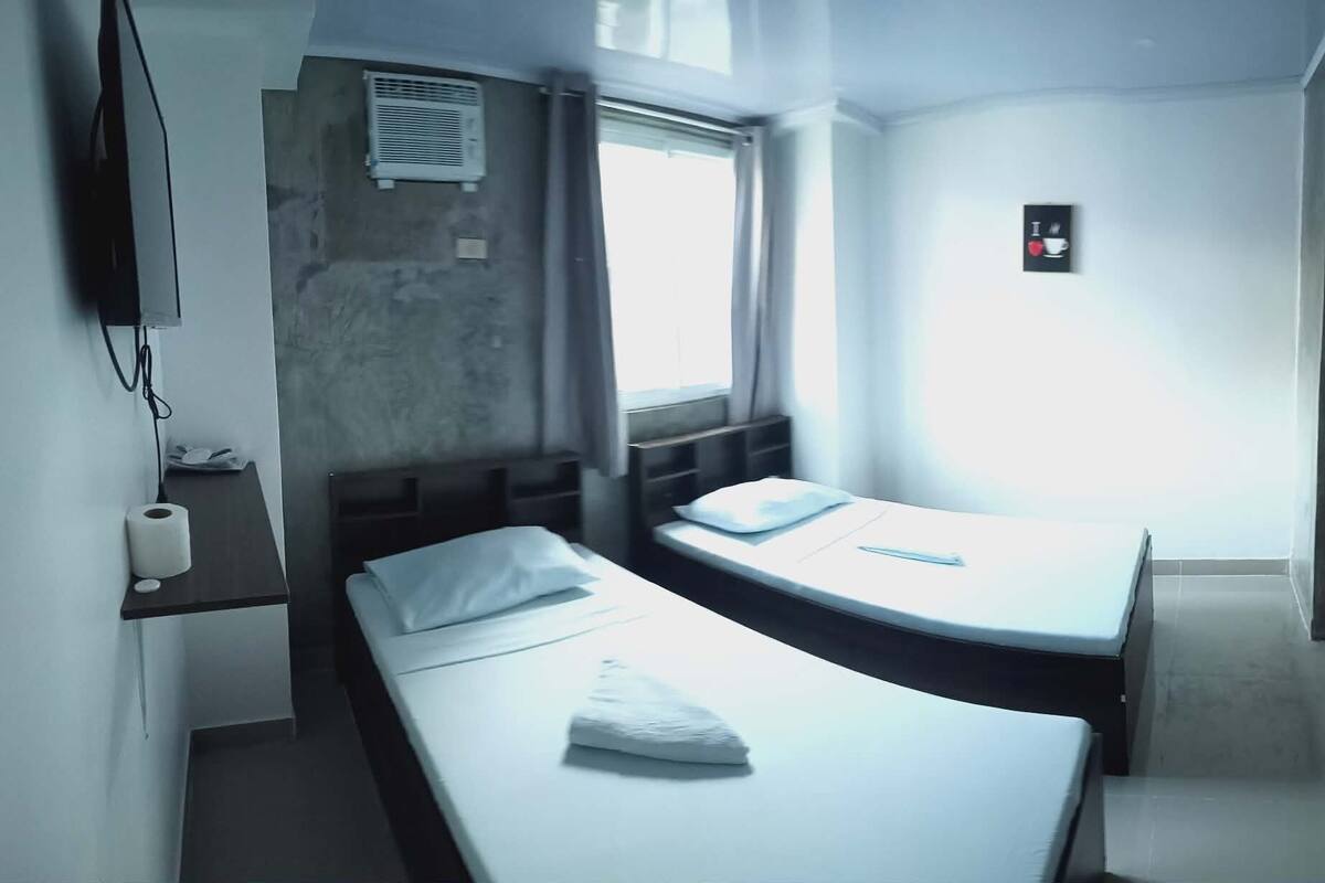 A small hotel room with two single beds, a window, a wall-mounted air conditioner, and a small shelf with a roll of toilet paper.