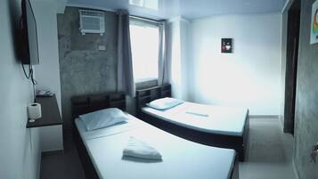 Une petite chambre d’hôtel avec deux lits simples, une fenêtre, un climatiseur fixé au mur et une petite étagère sur laquelle se trouve un rouleau de papier toilette.