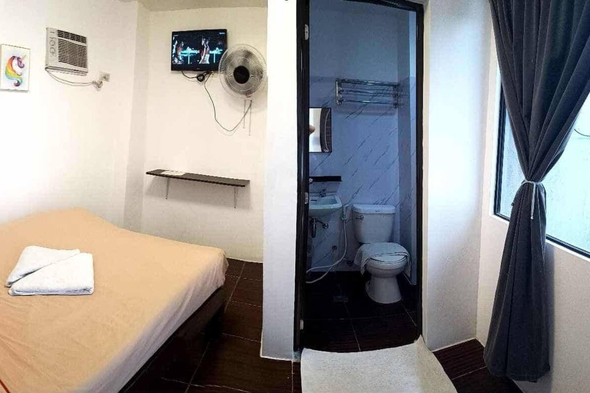 A room with a bed, a red pillow, a white towel, a wall-mounted TV, a fan, and a small shelf.