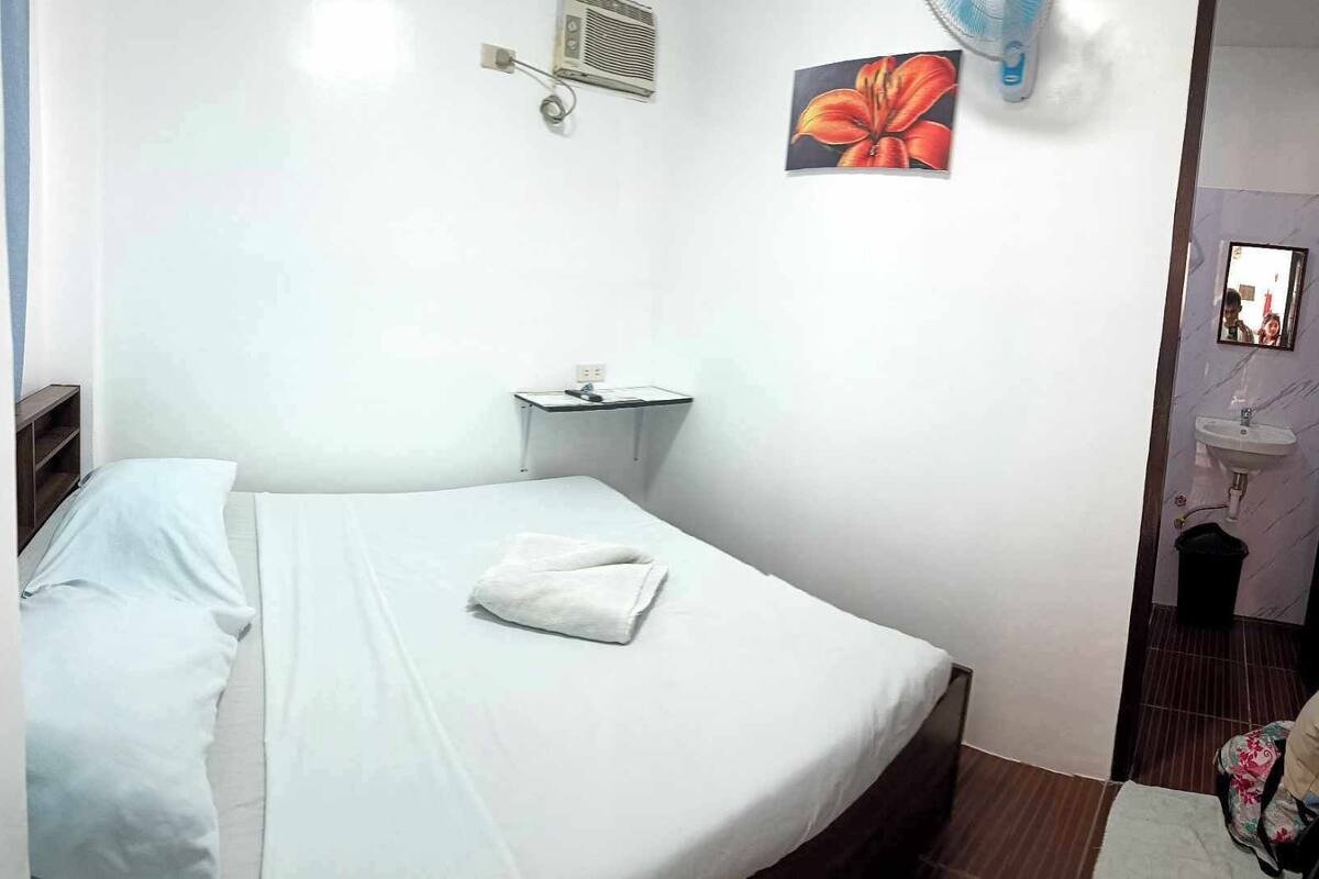 A small, clean room with a single bed, a bedside table, a wall-mounted air conditioner, a small sink, and a mirror.