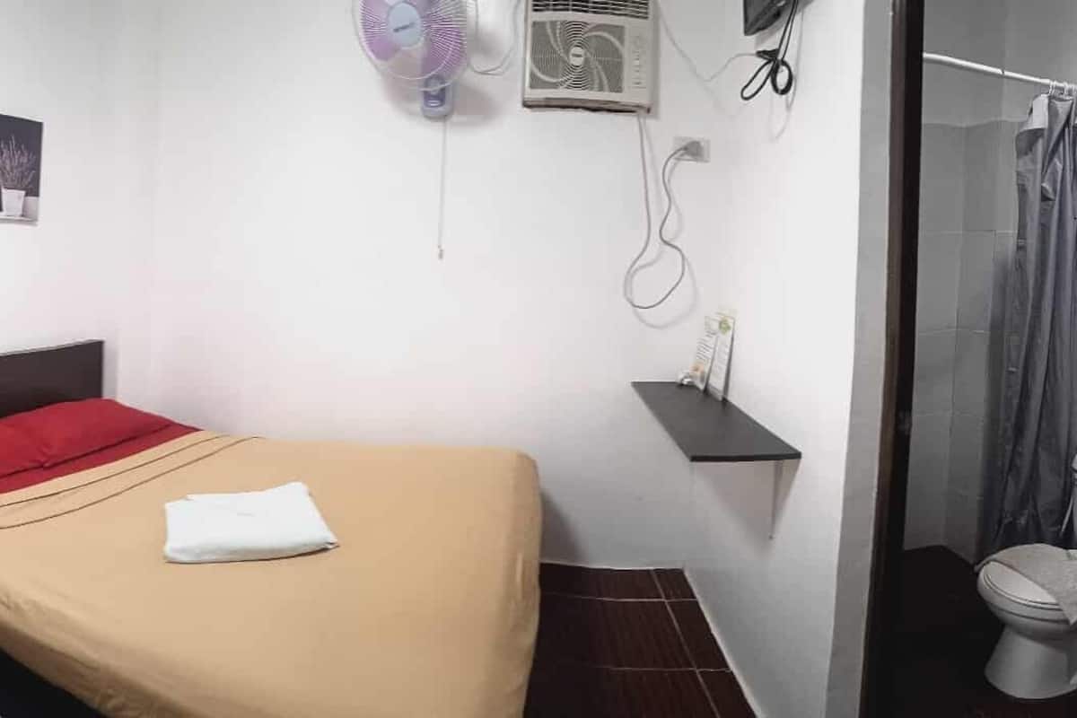 A compact hotel room with a single bed, a small bedside table, a wall-mounted fan, and a toilet.