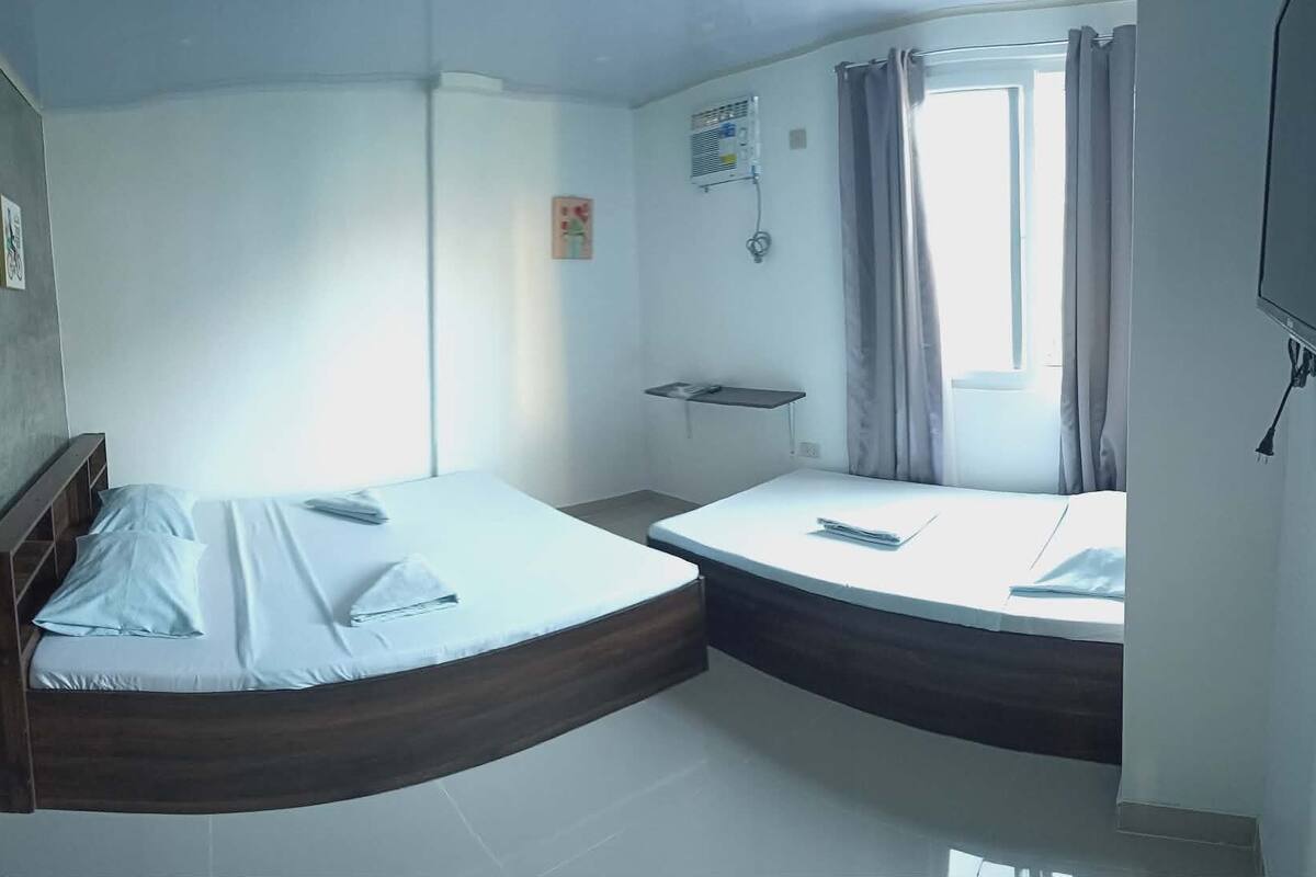 A small, modern room with two beds, a window with curtains, a flat-screen TV, and a small shelf.