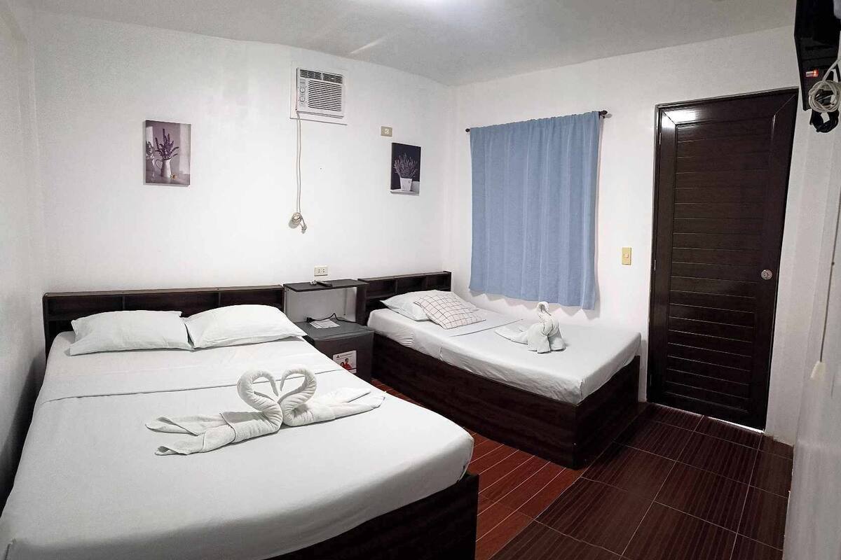 A hotel room with a large bed, a smaller bed, a nightstand, a desk, a window with a curtain, and a door.