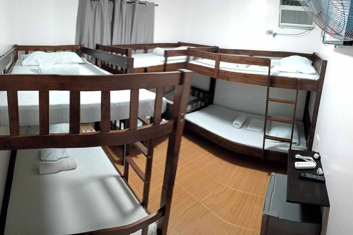 A dormitory room with bunk beds, a desk with a chair, a fan, and an air conditioning unit.