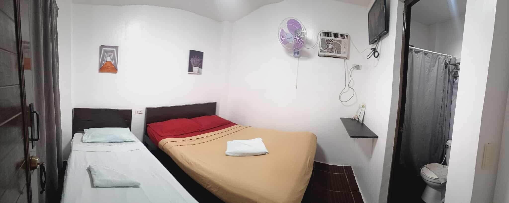 A small, compact hotel room with two single beds, a red and a beige bedspread, a wall-mounted air conditioner, a small shelf with a phone, and a toilet visible through an open door.