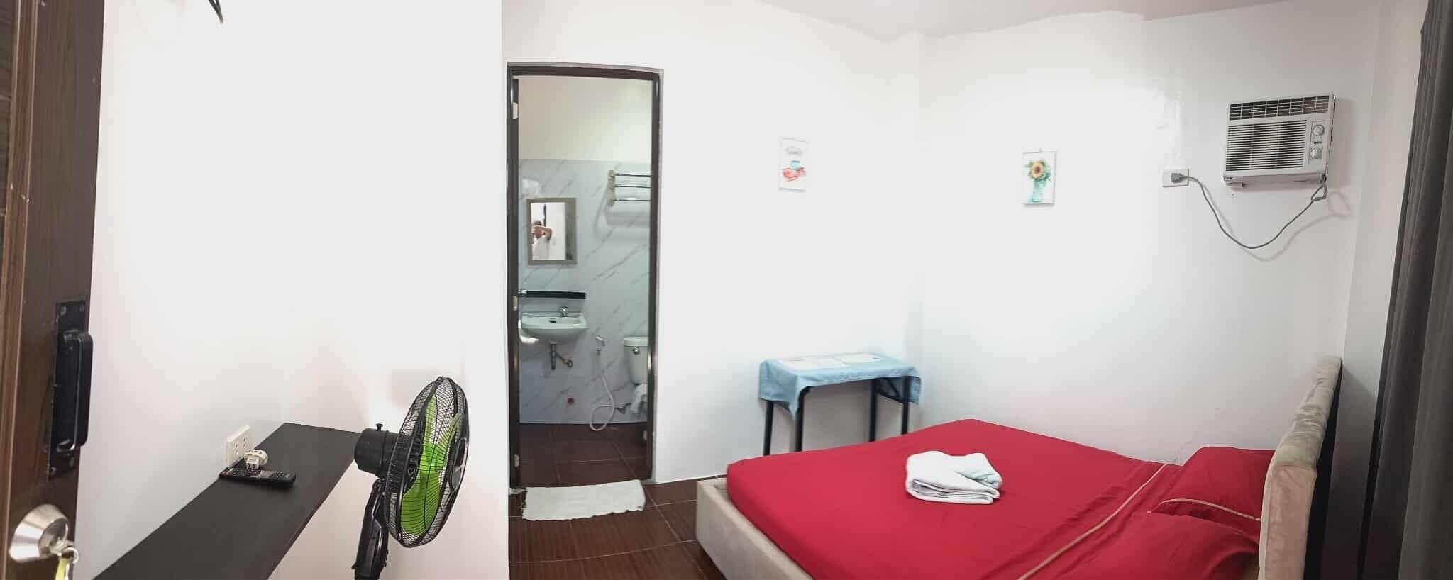 A hotel room with a bed, a desk with a fan, a bathroom with a sink and toilet, and an air conditioning unit.