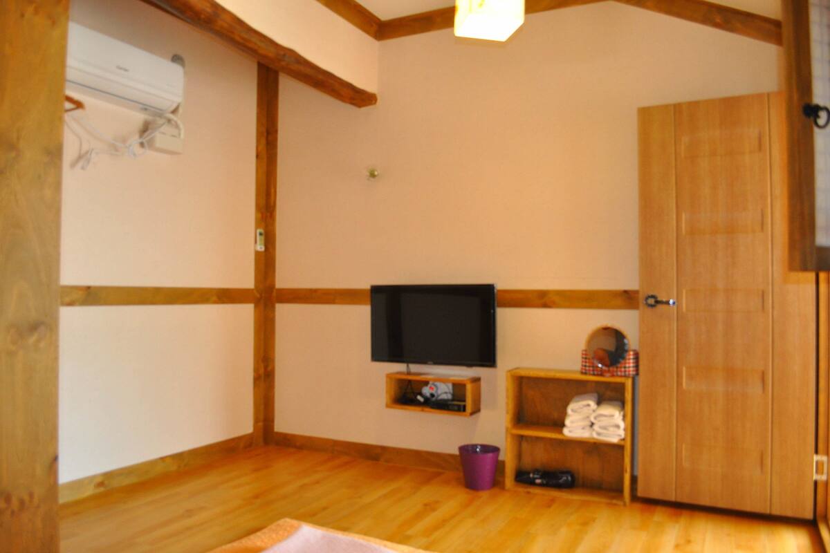 A room with wooden walls and floor, a television on a shelf, a door, and a small shelf with towels.
