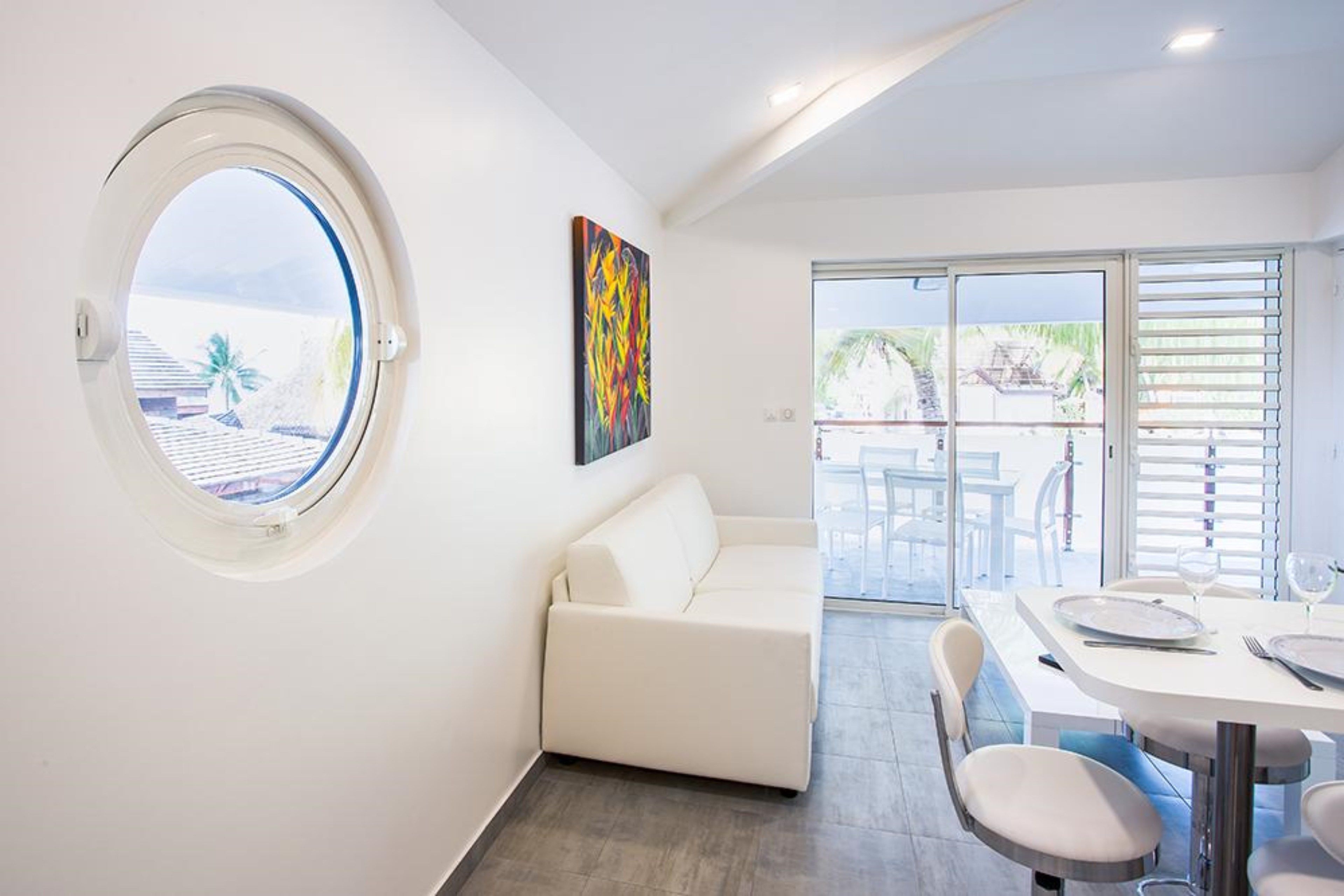 A modern room with a round window, a white sofa, a dining table with chairs, and a view of the outside.