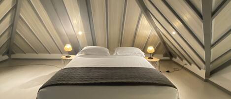 Een slaapkamer met een bed in het midden van de kamer, twee bedlampen en een schuin dak met zichtbare balken.