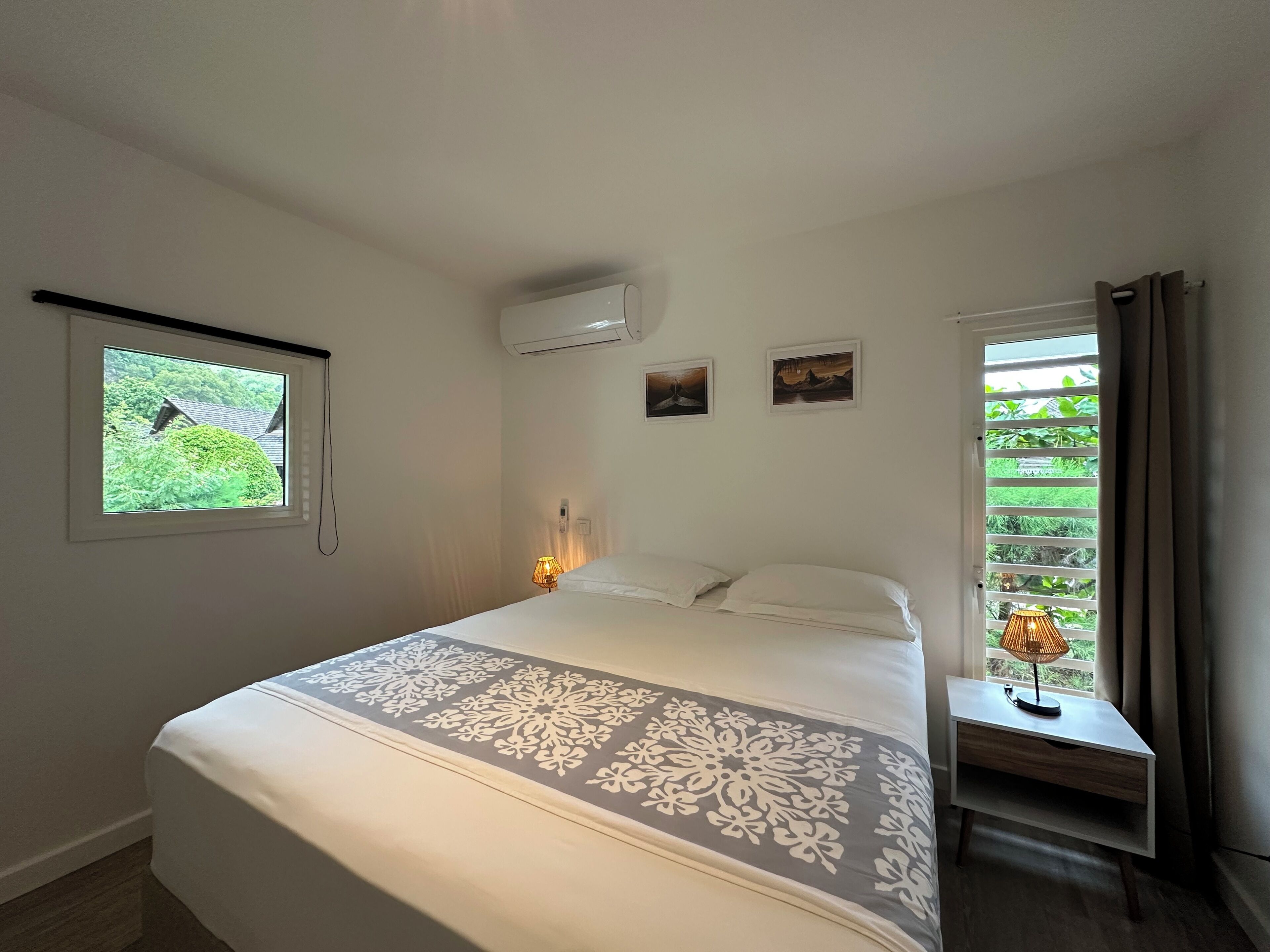 A bedroom with a bed, a window with a view of greenery, an air conditioner, and a bedside table with a lamp.