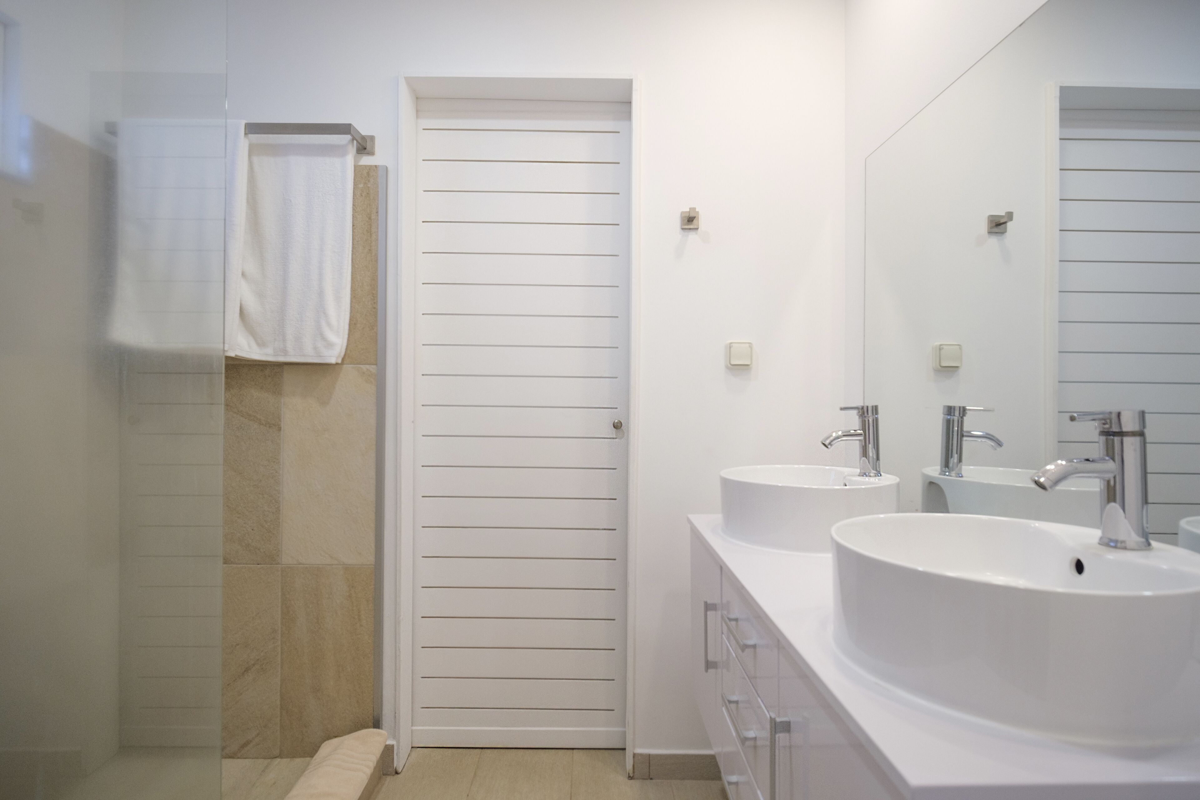 A modern bathroom with two sinks, a large mirror, and a door leading to a shower area.