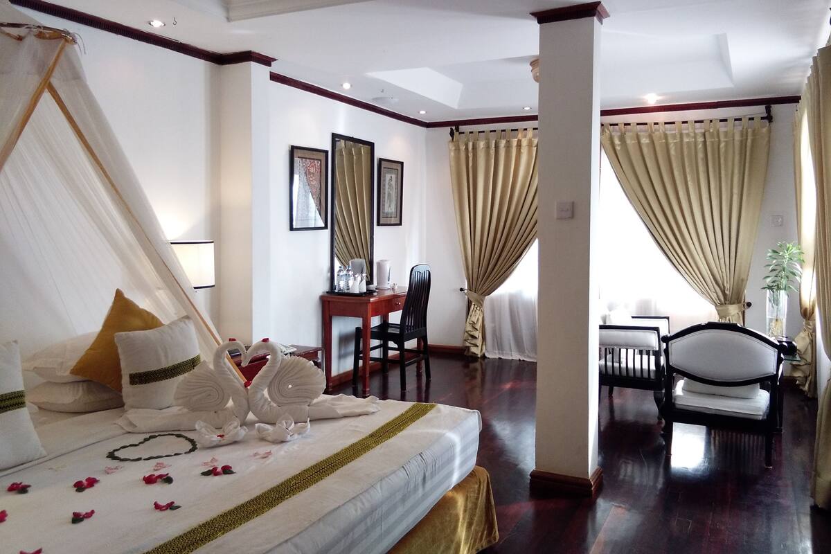 Une chambre d’hôtel avec un lit, un bureau et une chaise. Il y a des rideaux aux fenêtres et une plante sur le bureau.