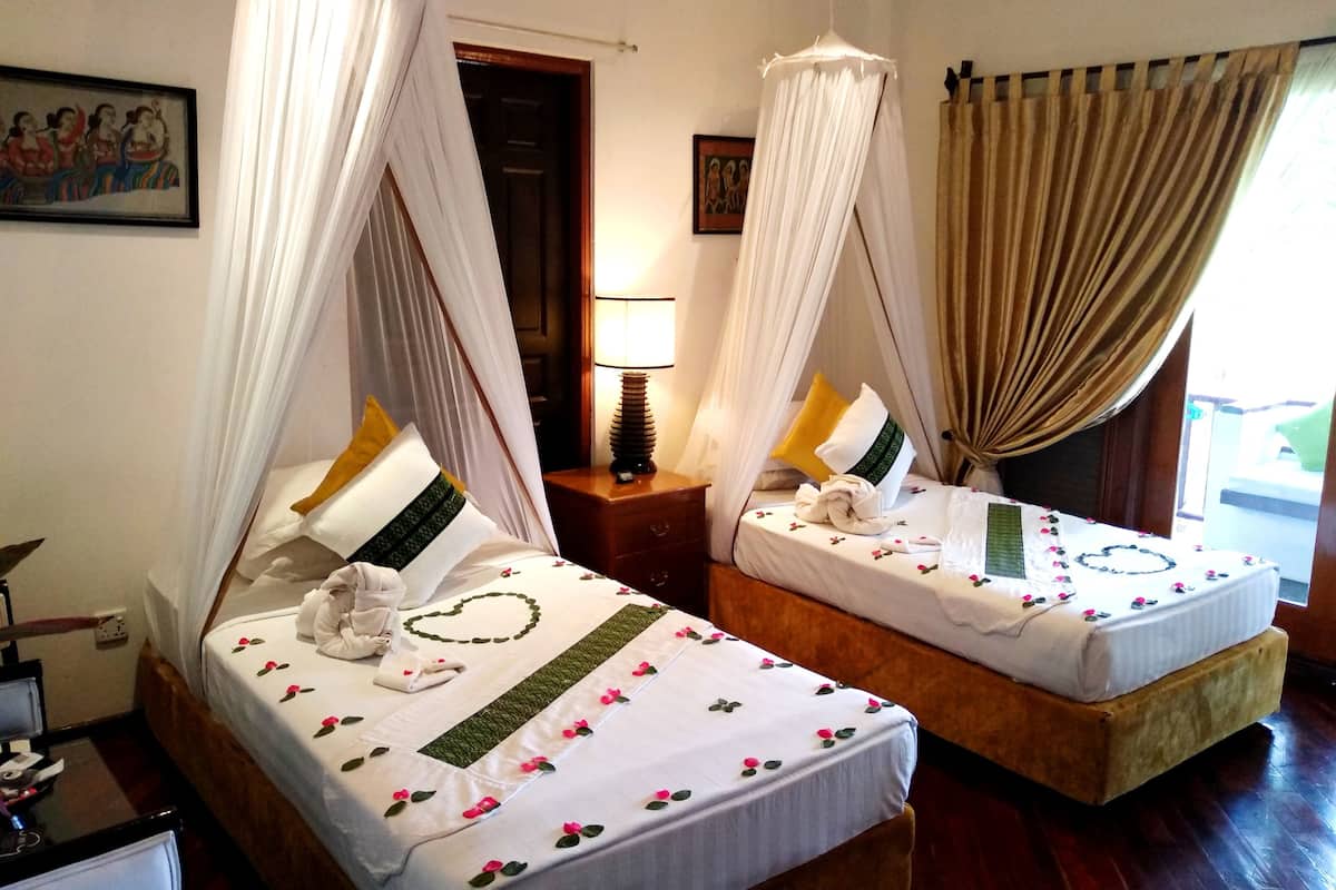 Deux lits doubles à baldaquin, du linge de lit blanc orné de motifs floraux, une table de chevet avec une lampe et un plancher en bois.