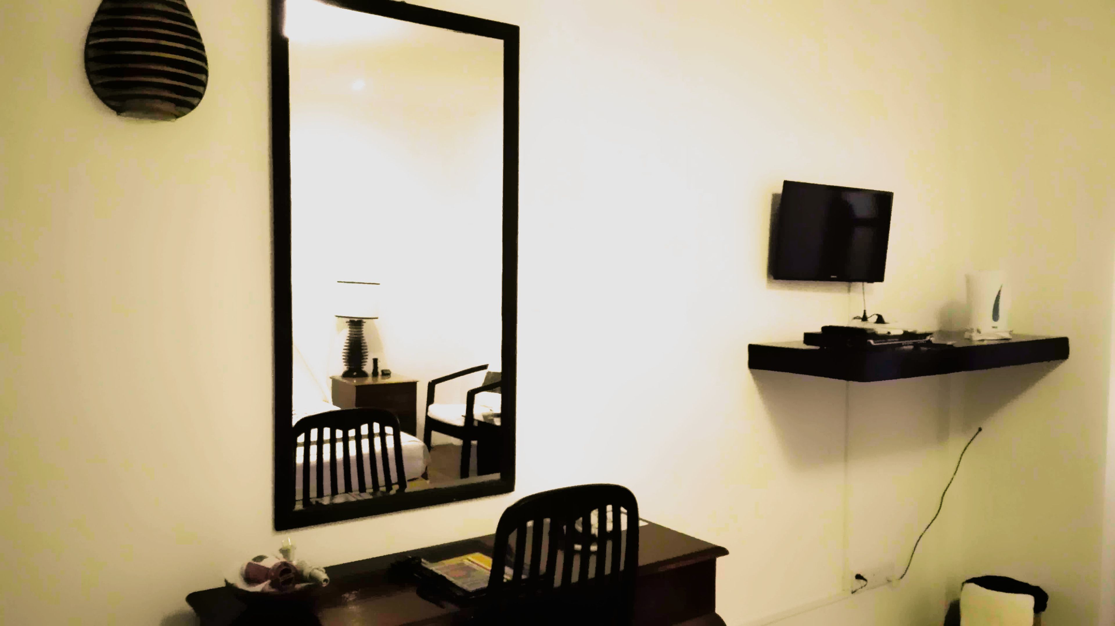 A room with a large mirror, a television on a shelf, a desk with a chair, and a lamp.