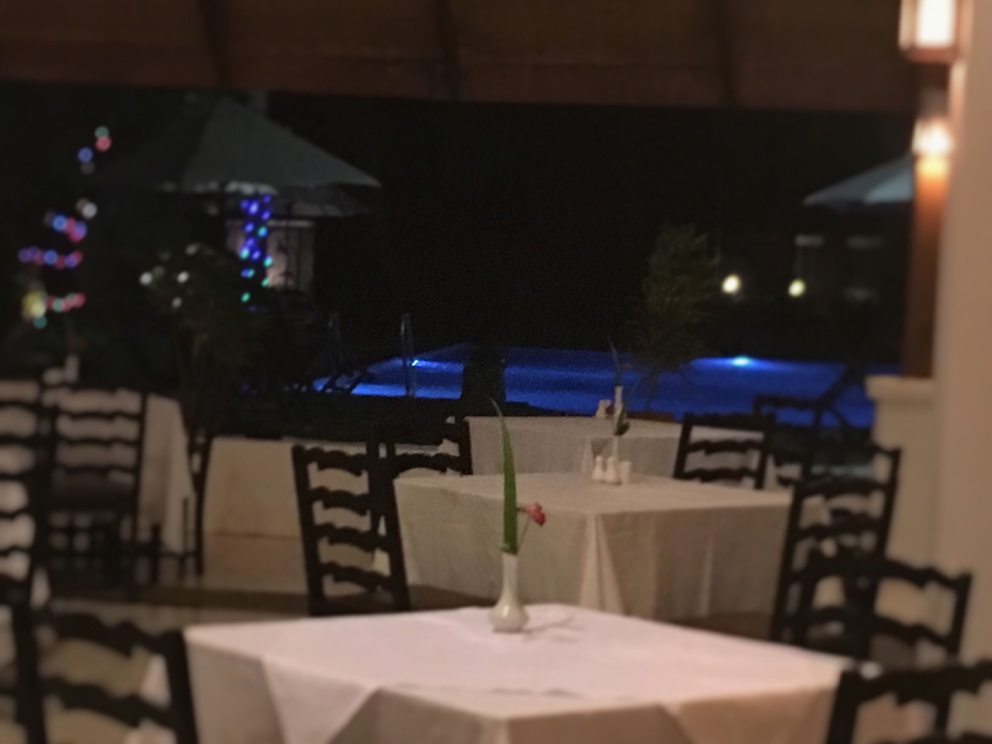 Un espace extérieur aménagé pour les repas, avec des tables et des chaises, une petite table ornée d’un vase et d’une fleur, et une vue sur une étendue d’eau la nuit.