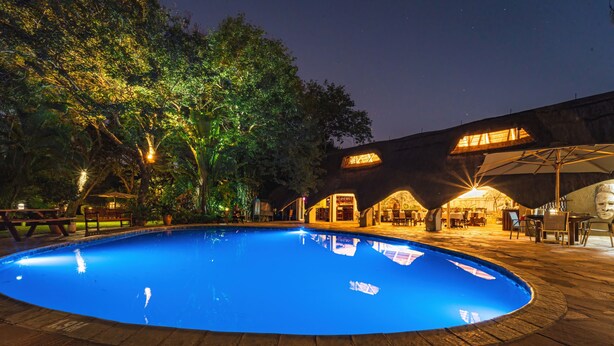 A swimming pool surrounded by a patio area with tables and chairs, and a building with multiple windows and a covered area in the background. - Bayete Guest Lodge (Victoria Falls)
