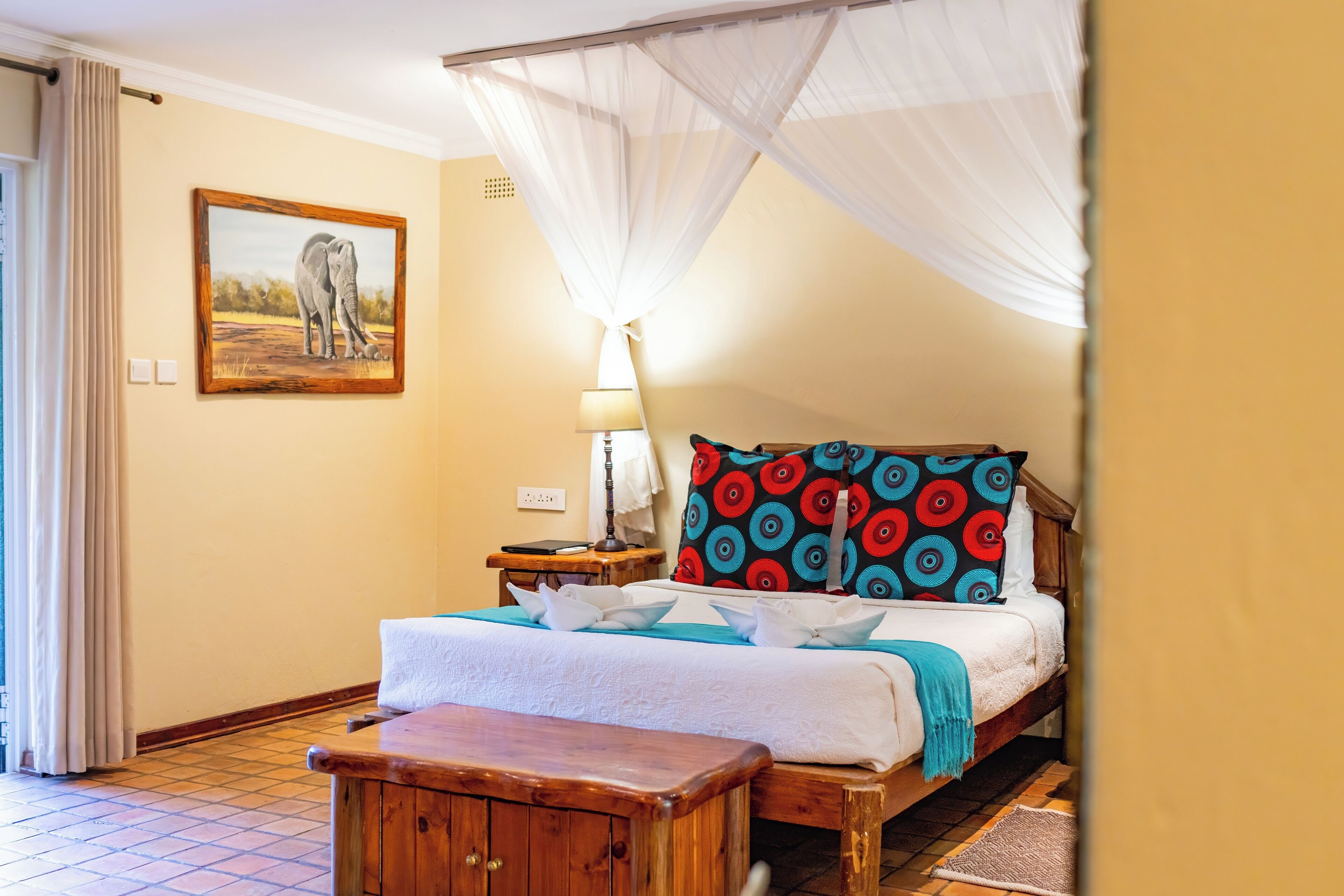 A bedroom with a bed, a nightstand, a lamp, a wooden bench, a mosquito net, and a painting on the wall.