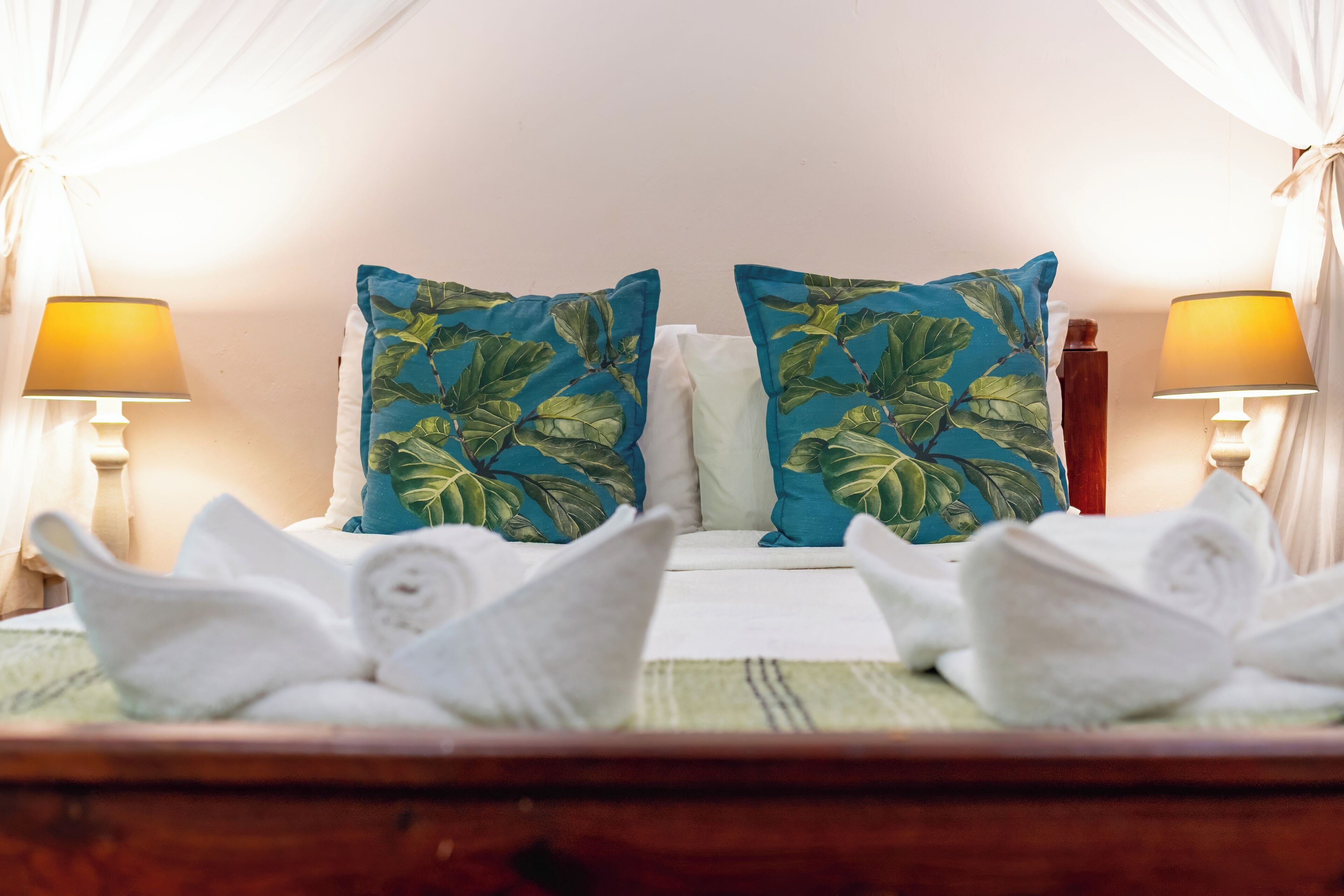A neatly made bed with two pillows featuring a tropical leaf pattern, white towels, and two bedside lamps with yellow shades.