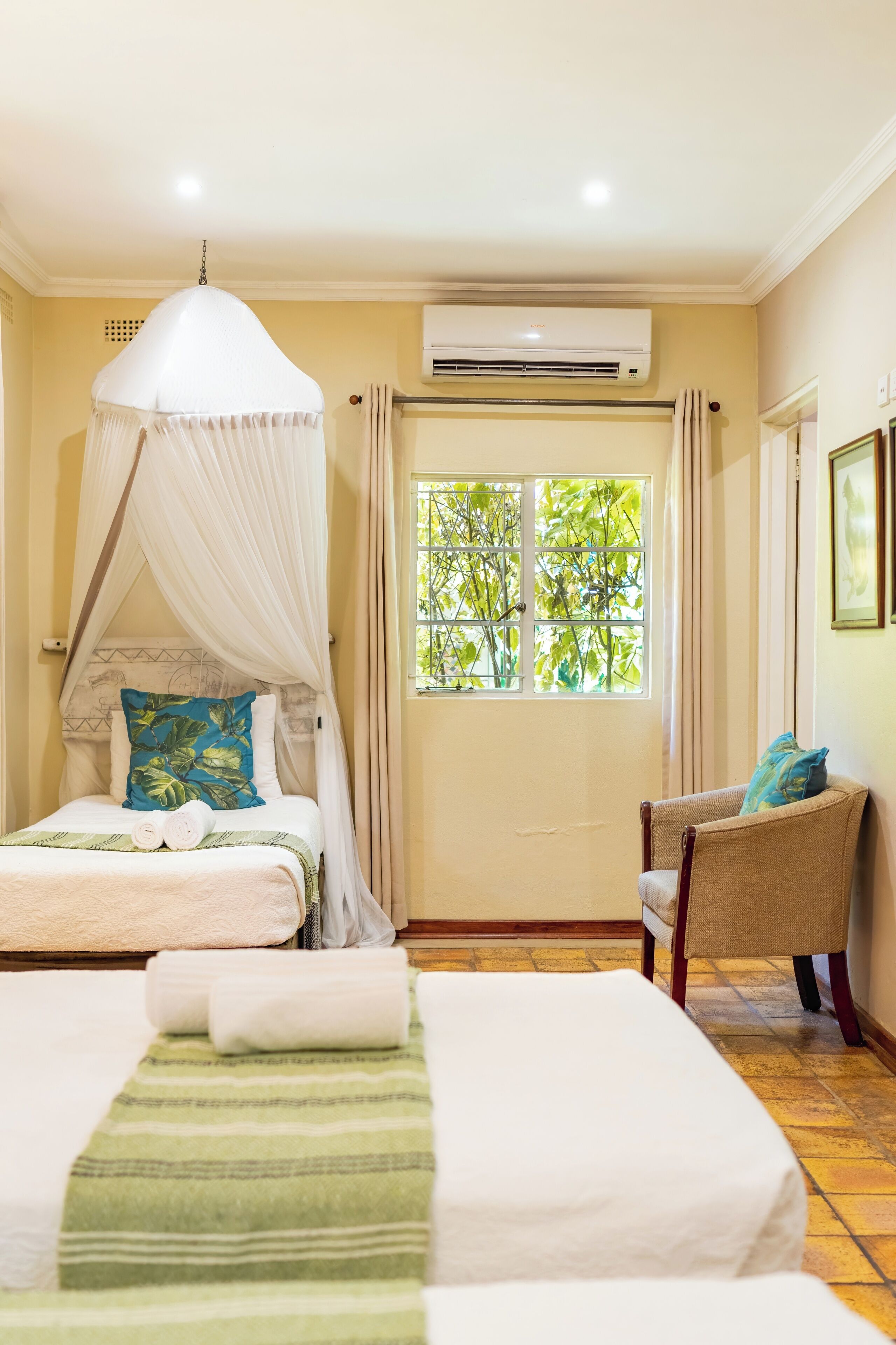A bedroom with two beds, a canopy bed, a chair, an air conditioning unit, and a window with a view of trees.