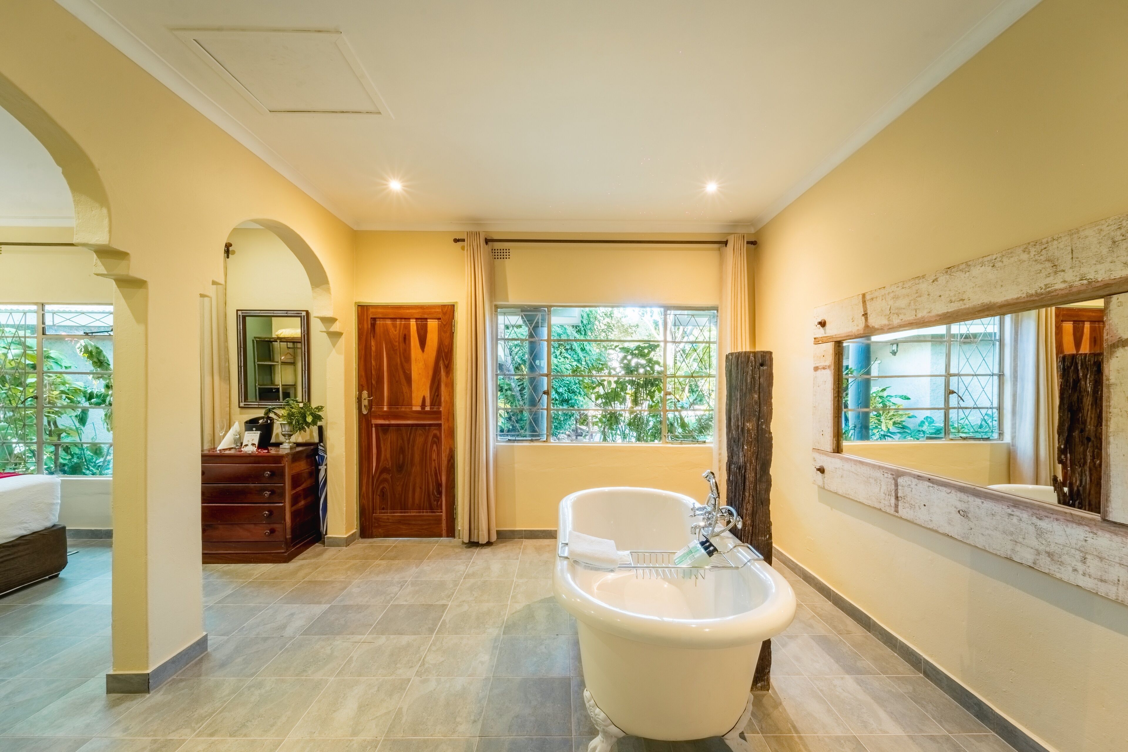 A bathroom with a freestanding bathtub, wooden cabinets, a mirror, and large windows with a view of greenery.