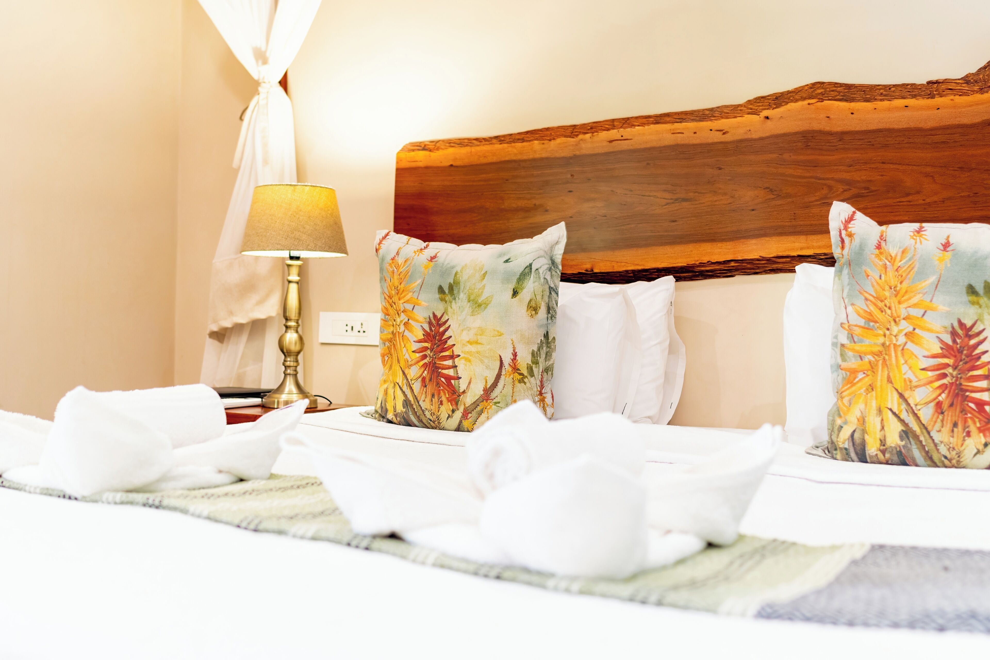 A neatly made bed with floral pillows, a bedside lamp, and a white curtain.