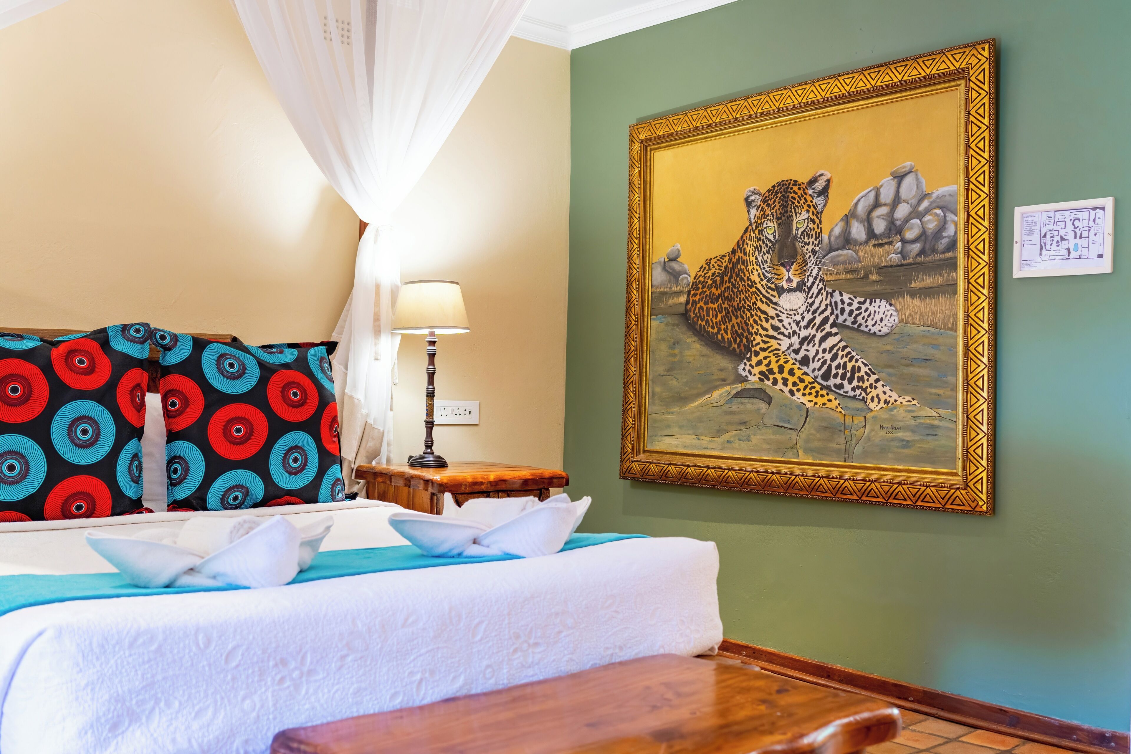 A hotel room with a bed, a nightstand, a lamp, and a painting of a leopard and a bird on the wall.