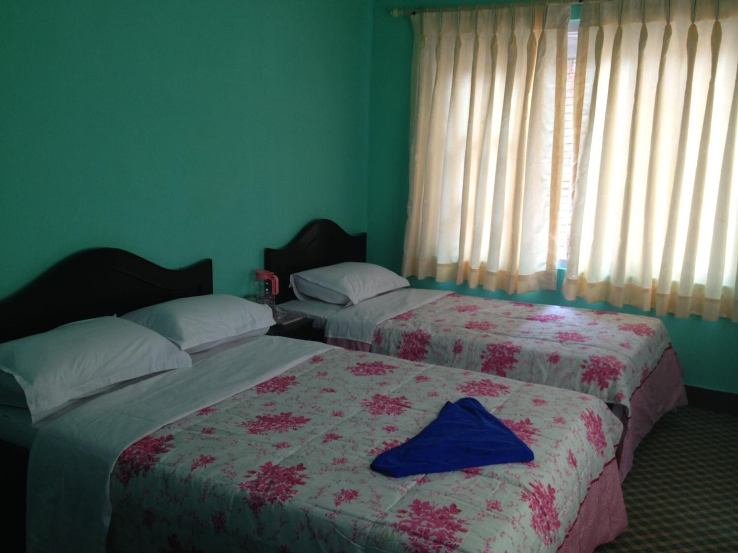 Quarto com duas camas, cada uma com roupa de cama florida, uma mesa de cabeceira com abajur e uma janela com cortinas leves.