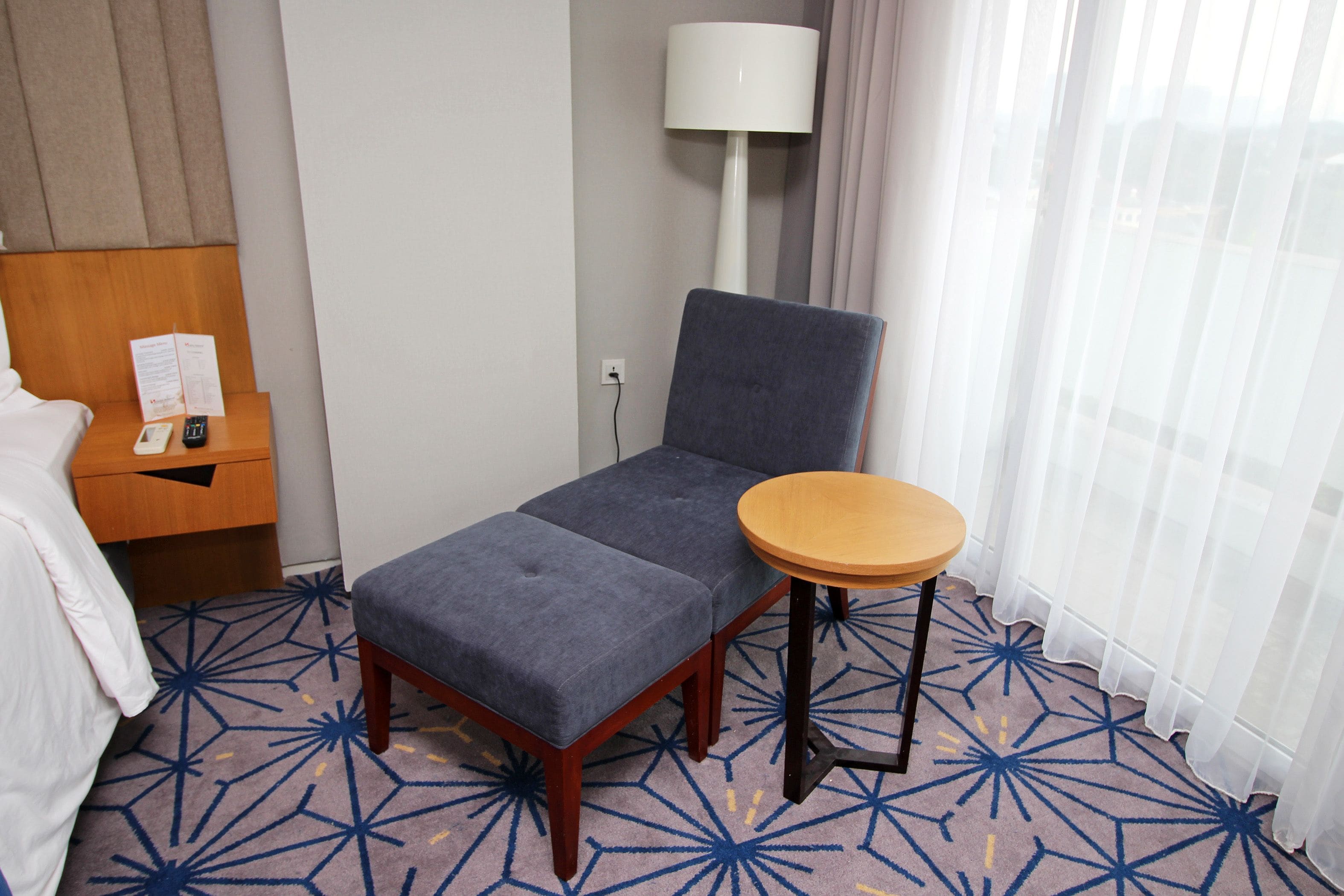 A hotel room with a bed, a small desk with a lamp, a chair, and a round table.
