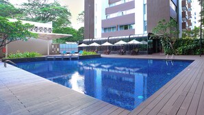 Una piscina con sillones de descanso y sombrillas, rodeada por un edificio con balcones y una zona de estar techada.