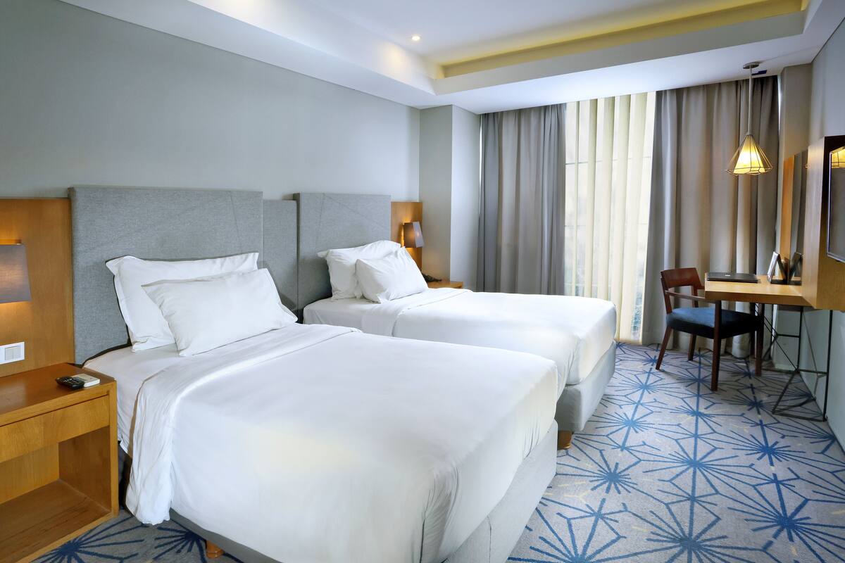 A hotel room with two beds, a desk, a chair, and a large window with curtains.