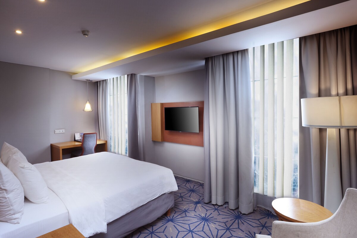 Habitación de hotel con una cama grande, un escritorio, una silla, un televisor y una ventana con cortinas.