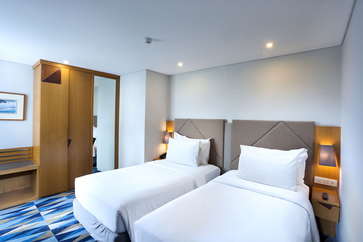 Habitación de hotel con dos camas, un armario de madera, un banco y una lámpara de pared.