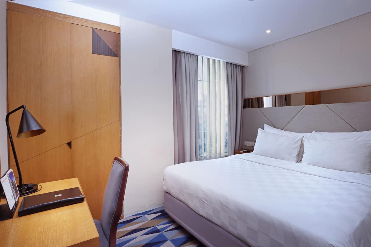 A hotel room with a bed, a desk with a laptop, a chair, and a large window with curtains.