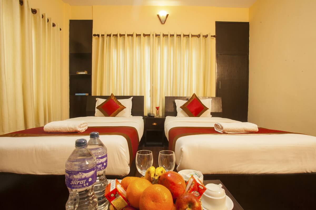 Habitación de hotel con dos camas, una mesita de noche, una mesa con frutas y agua embotellada, y una cortina.