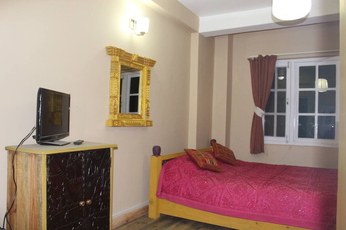 A bedroom with a bed, a TV on a wooden cabinet, a mirror, and a window with curtains.