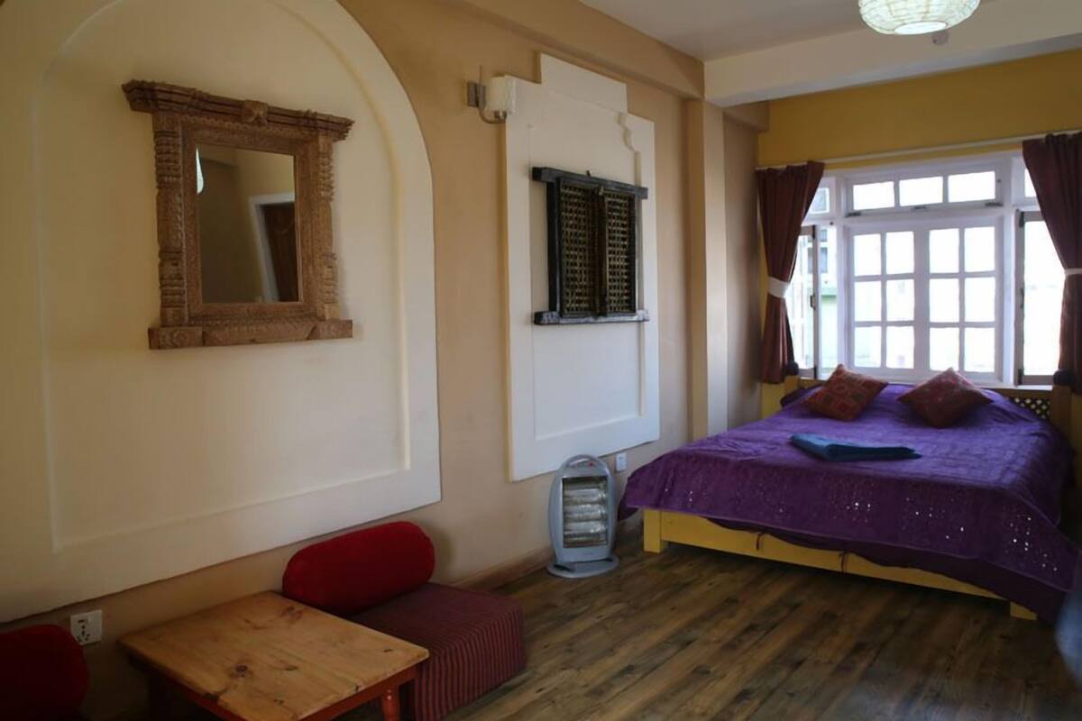 A room with a bed, a table, a bench, a heater, and a window with curtains.