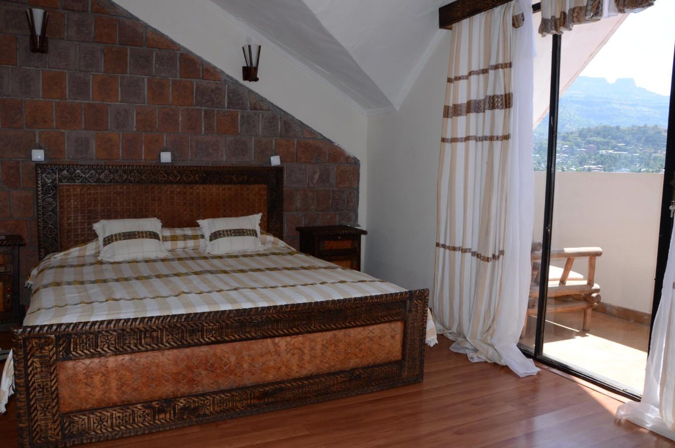 A bedroom with a large bed, a brick wall, a wooden floor, a nightstand, and a view of the outdoors.