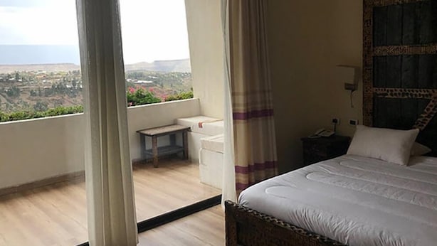 A bedroom with a large bed, a nightstand, a small table, and a view of a landscape through a window. - Lalibela Lodge (Lalibela)