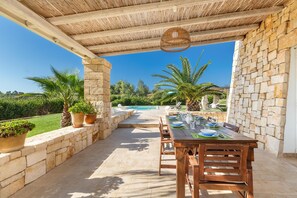 Eine Terrasse im Freien mit einem Holztisch, der zum Essen gedeckt ist, Steinmauern und Blick auf einen Swimmingpool und Grünflächen.