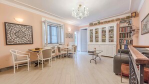 A spacious living room with a chandelier, a dining area with white chairs, a bookshelf, and a staircase. - B&B Palazzo Benso (Palermo)