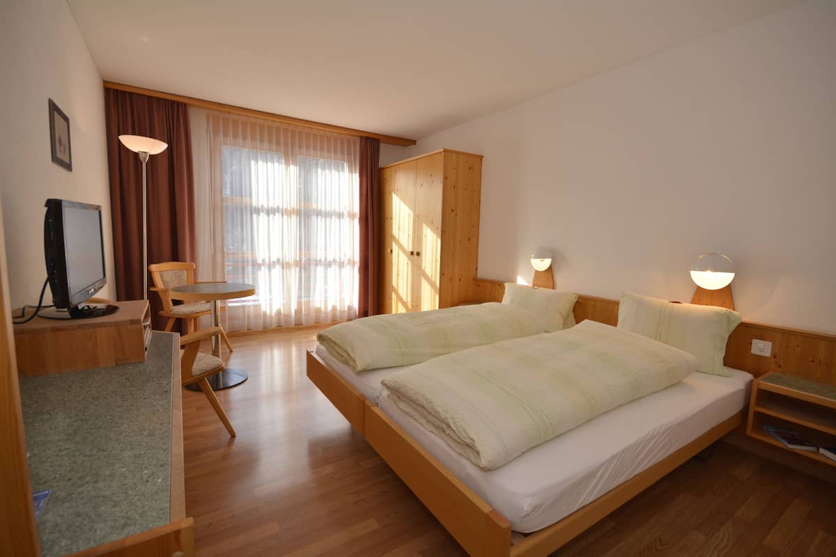 Habitación de hotel con una cama de madera, un escritorio con ordenador, una silla y un amplio ventanal con cortinas.
