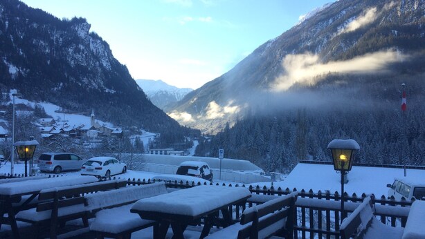 A snow-covered outdoor seating area with benches and tables, a parking sign, and a mountain backdrop.