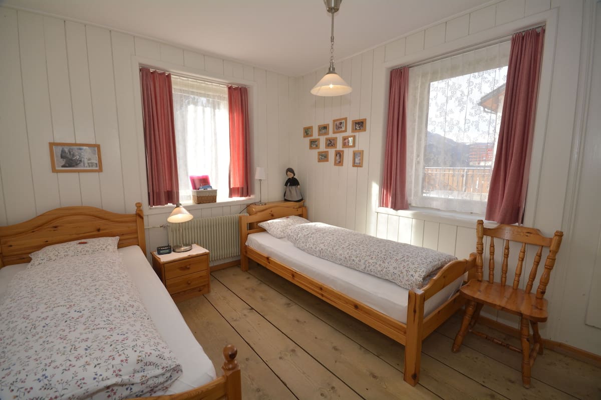 Un dormitorio con dos camas de madera, una silla de madera, una mesita de noche, una lámpara y una ventana con cortinas.